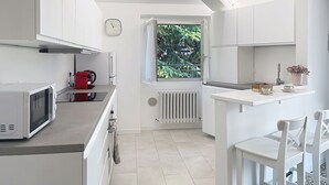 Ett modernt kök med vita skåp, en mikrovågsugn, en röd vattenkokare och ett fönster med utsikt över grönområden.