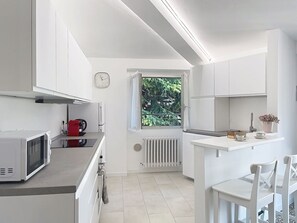 Eine moderne Küche mit weißen Schränken, einer Mikrowelle, einem roten Wasserkocher und einem Fenster mit Blick auf Grünflächen.