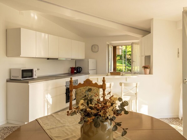 Een moderne keuken met witte kastjes, een magnetron en een koffiezetapparaat. Er is een eethoek met een houten stoel en een vaas met bloemen.