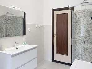Ein Badezimmer mit weißem Waschbecken, einem Spiegel, einer Duschkabine und einer Tür mit Holzverkleidung.