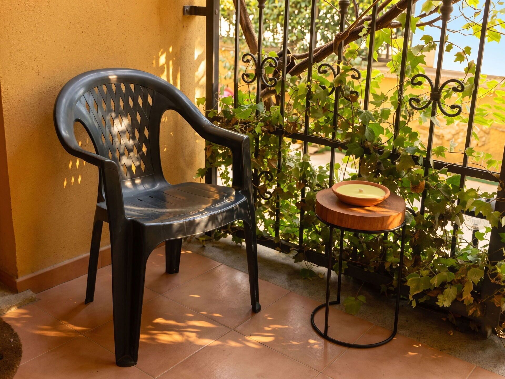 Une chaise en plastique noire, posée sur un sol carrelé, à côté d’une clôture en fer forgé ornée de plantes grimpantes.