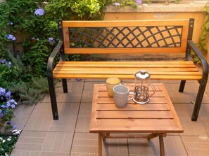 A wooden bench with a metal lattice backrest, a wooden table with a mug and a thermos, and a garden with purple flowers. - Wonderful  villa for 4 guests with WIFI, A/C, TV and patio (Olbia)