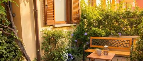 A courtyard with a wooden bench, a table with a cup and a lantern, and a variety of plants and flowers.