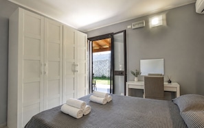 Ein Schlafzimmer mit Bett, Schrank, Badezimmerspiegel, Nachttisch mit Vase und Blick ins Freie.