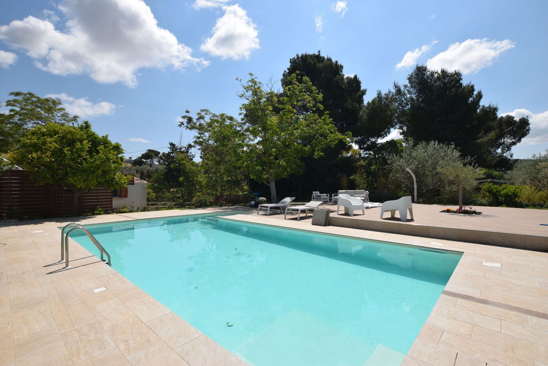 Una piscina rectangular con un borde de baldosas, rodeada de árboles y mobiliario exterior.