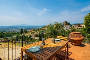 Ein Holztisch mit Tellern, einer Tasse und einer Kaffeekanne auf einer Terrasse im Freien mit Blick auf eine Stadt und Berge.