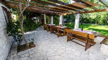 Een terras buiten met stenen vloer, houten tafels en banken, en een pergola.