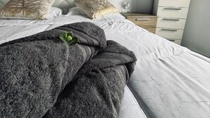 Siististi pedattu sänky, jossa on harmaa peitto ja kaksi koristeellista tyynyä, valkoinen lipasto ja pieni ruukkukasvi yöpöydällä.