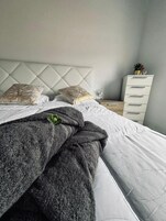 Una cama bien tendida con un edredón gris y dos almohadas decorativas, un aparador blanco y una pequeña planta en maceta sobre una mesita de noche.