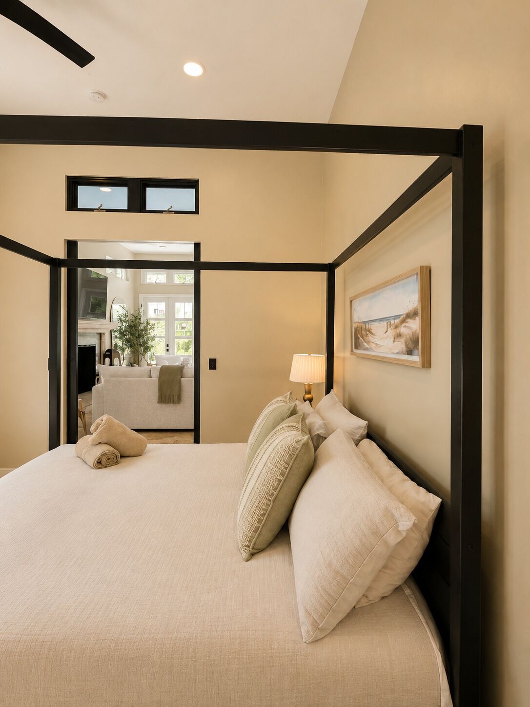 A four-poster bed with a beige headboard and pillows, a bedside lamp, and a painting on the wall.