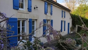 Ein zweistöckiges Gebäude mit blauen Fensterläden, einem Ziegeldach und einem Garten mit violetten Blumen.