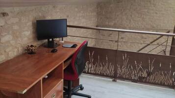 A room with a wooden desk, a computer, and a chair. There is a stone wall and a metal railing in the background.