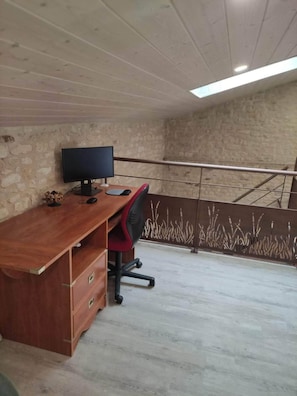 A room with a wooden desk, a computer, and a chair. There is a stone wall and a metal railing in the background. - Gîte les glycines (Saint-Sigismond)