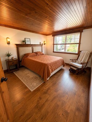 A bedroom with a wooden ceiling, a bed with a brown bedspread, a nightstand, a chair, and a window with a view of greenery.