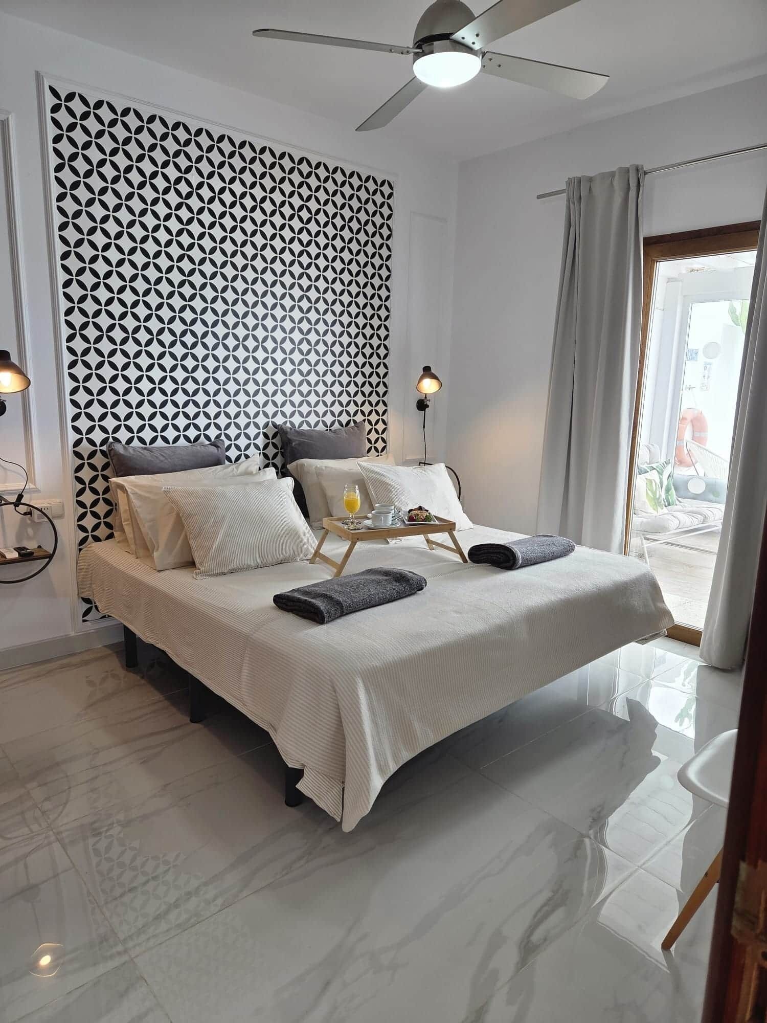 Une chambre d’hôtel moderne avec un grand lit, un ventilateur de plafond, une table de chevet avec un plateau, et une vue sur l’extérieur.