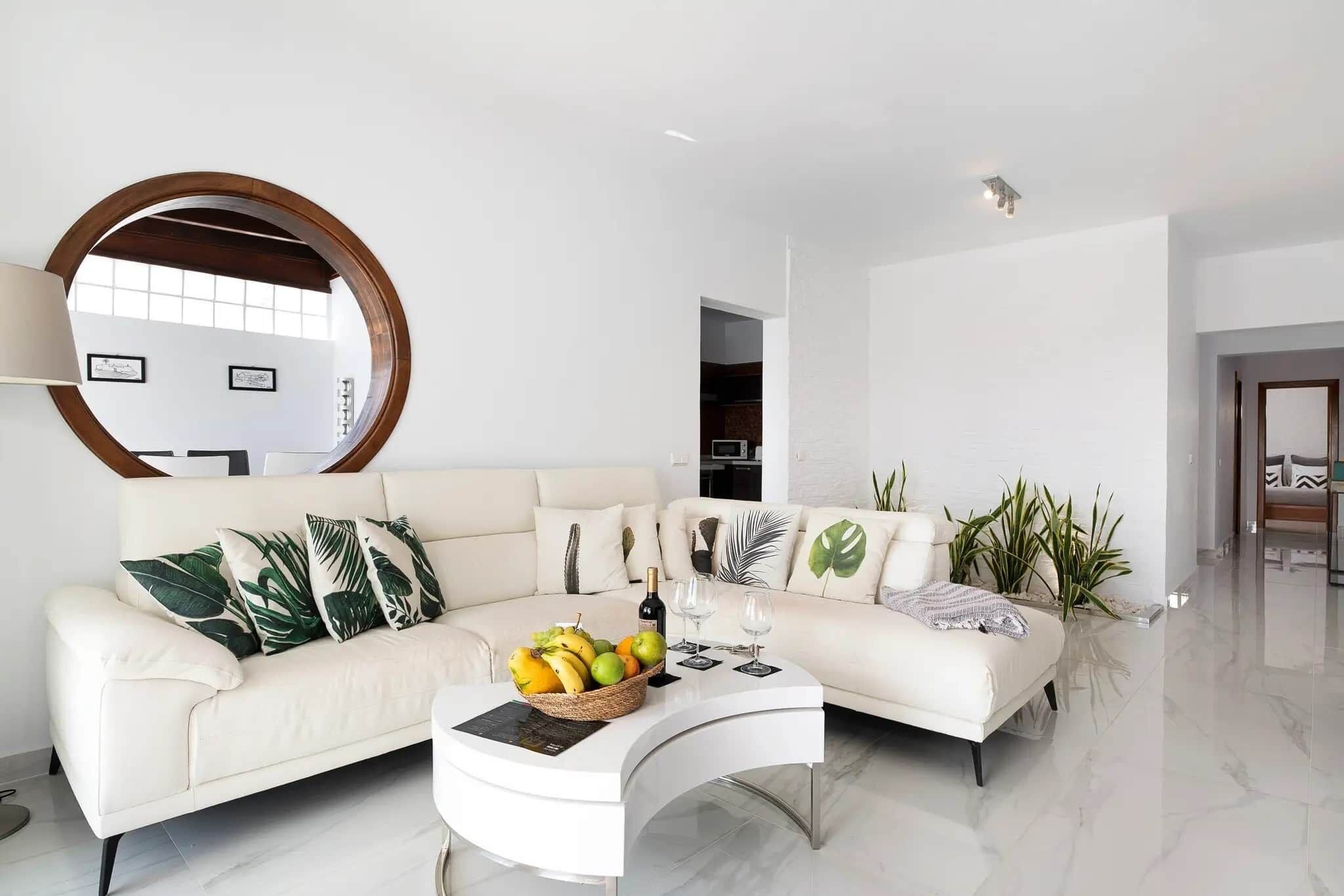 Un salon moderne avec un canapé blanc, un miroir rond et une table basse sur laquelle sont disposés des fruits.