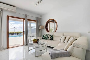 A modern living room with a white sofa, a round coffee table with fruits and wine glasses, and a view of a pool outside. - El Ancla - Luxury Villa with great View (Playa Blanca)