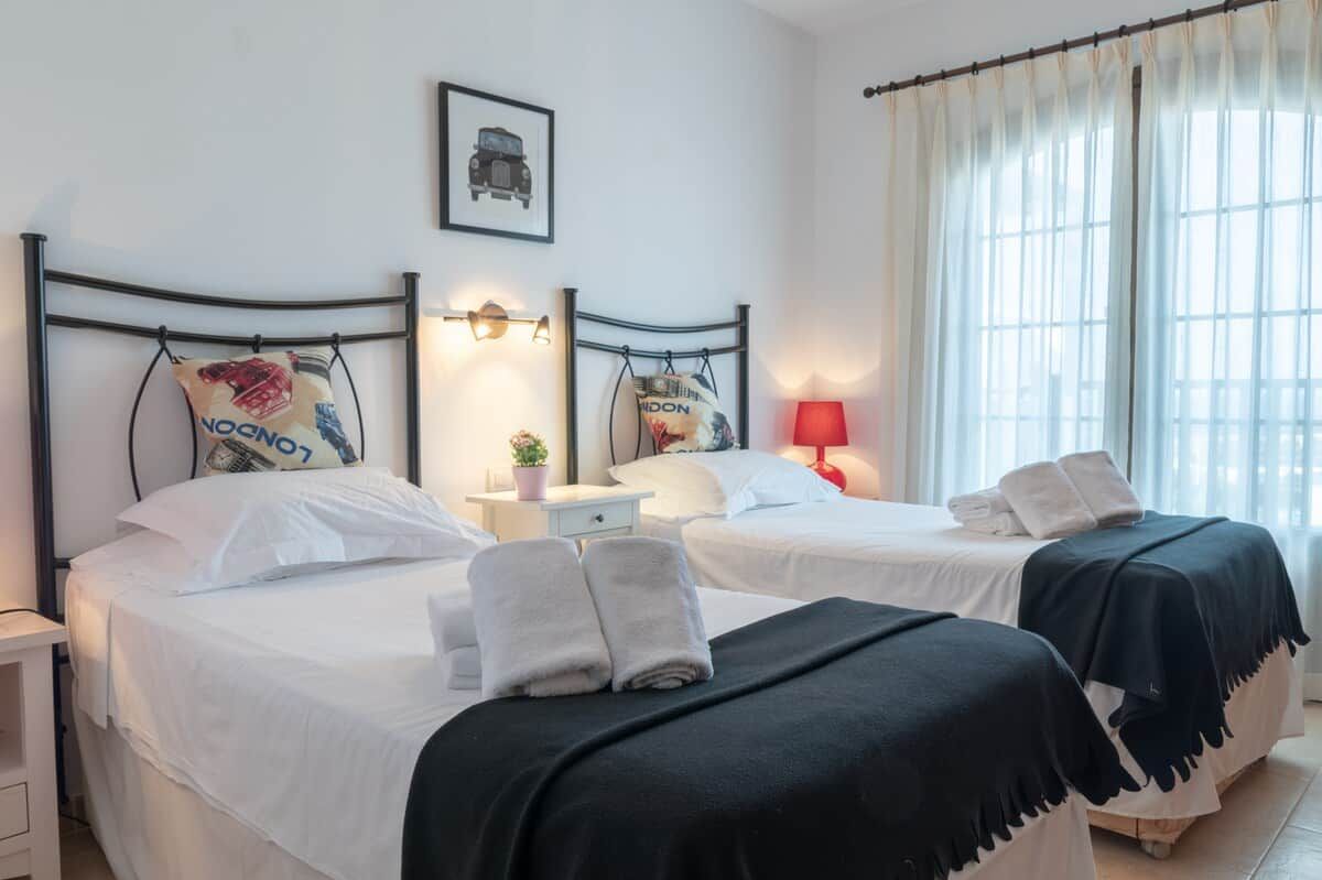 A hotel room with two beds, each with a pillow and a towel. There is a nightstand with a lamp and a small table with a plant. The room has a window with curtains and a framed picture on the wall.