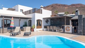 A swimming pool with lounge chairs and a barbecue area, surrounded by a white building with a mountain backdrop. - Luxury villa with a stunning sea view (Playa Blanca)