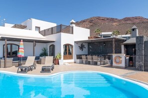 A swimming pool with lounge chairs and a barbecue area, surrounded by a white building with a mountain backdrop. - Luxury villa with a stunning sea view (Playa Blanca)