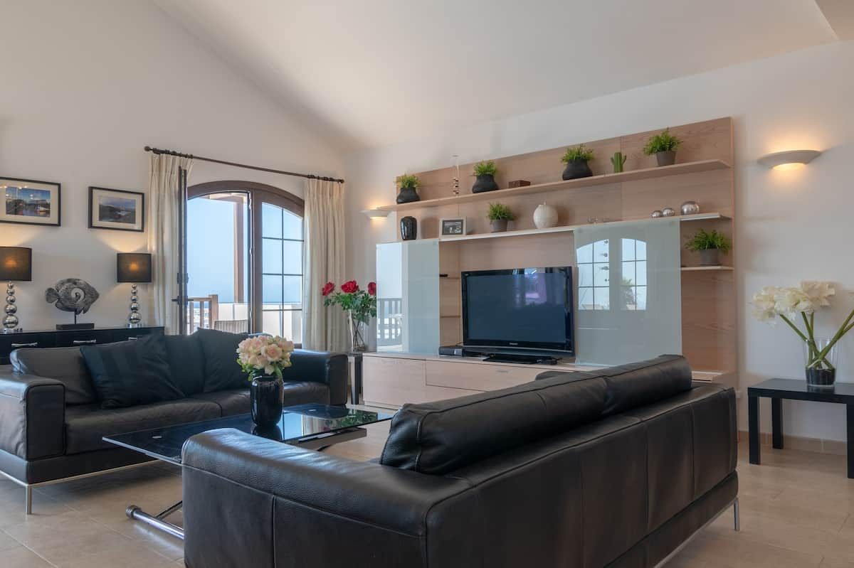 A modern living room with a sofa, coffee table, and a flat-screen TV. There are decorative plants and a vase with flowers on the shelves.