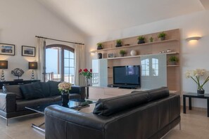A modern living room with a sofa, coffee table, and a flat-screen TV. There are decorative plants and a vase with flowers on the shelves. - Luxury villa with a stunning sea view (Playa Blanca)