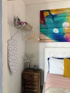 Ein Schlafzimmer mit einem Bett, einem Nachttisch, einem an der Wand befestigten Regal und einem farbenfrohen, abstrakten Gemälde.