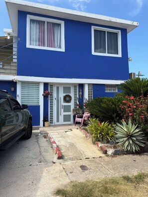 A two-story building with a blue exterior, a garage door, and a driveway leading to a front porch with a bench and potted plants.