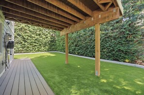 Una terraza de madera con una pérgola, rodeada de césped cuidado y follaje denso.