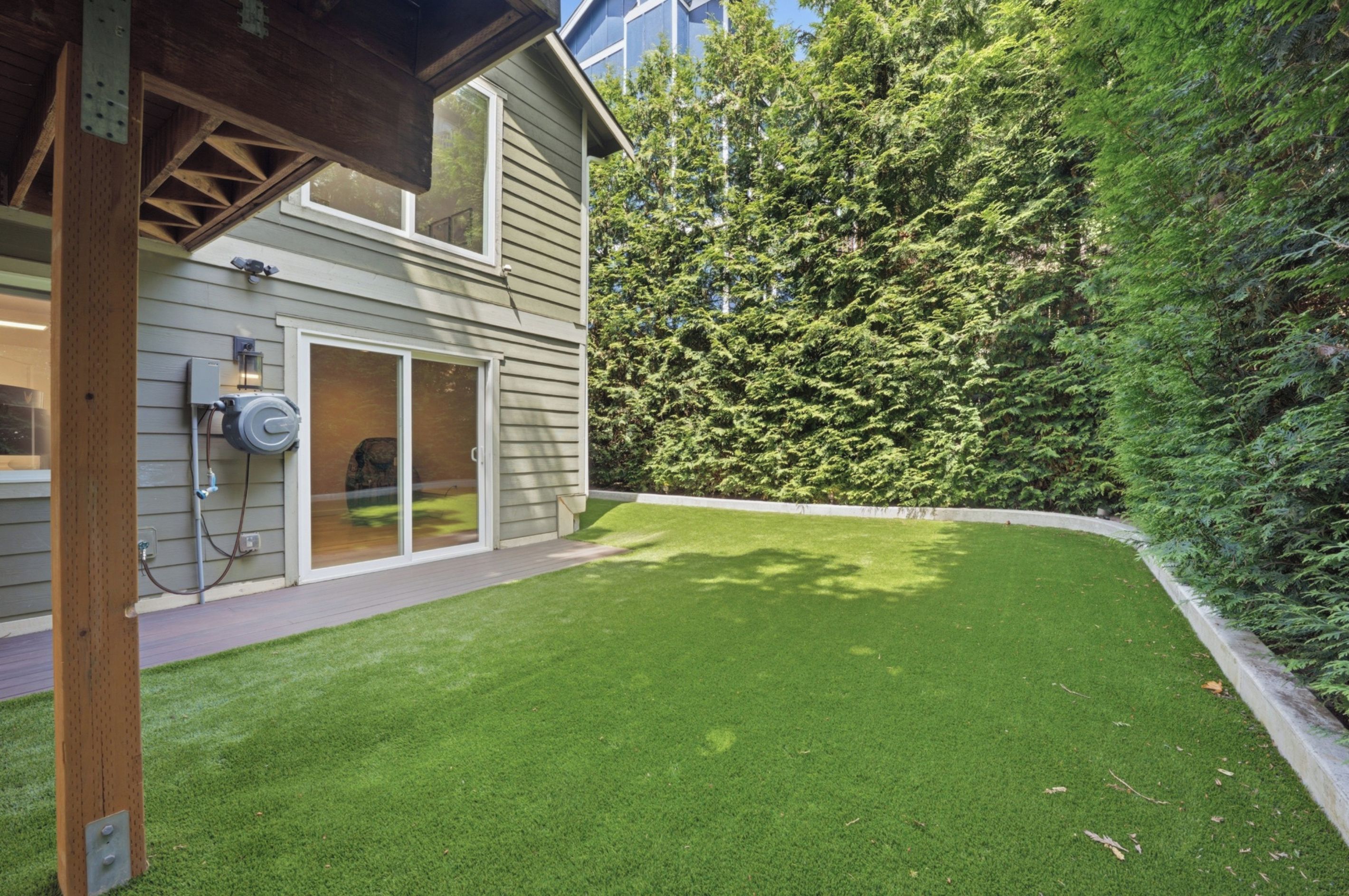 A residential property with a well-maintained lawn, a sliding glass door leading to a covered patio, and a dense evergreen forest in the background.