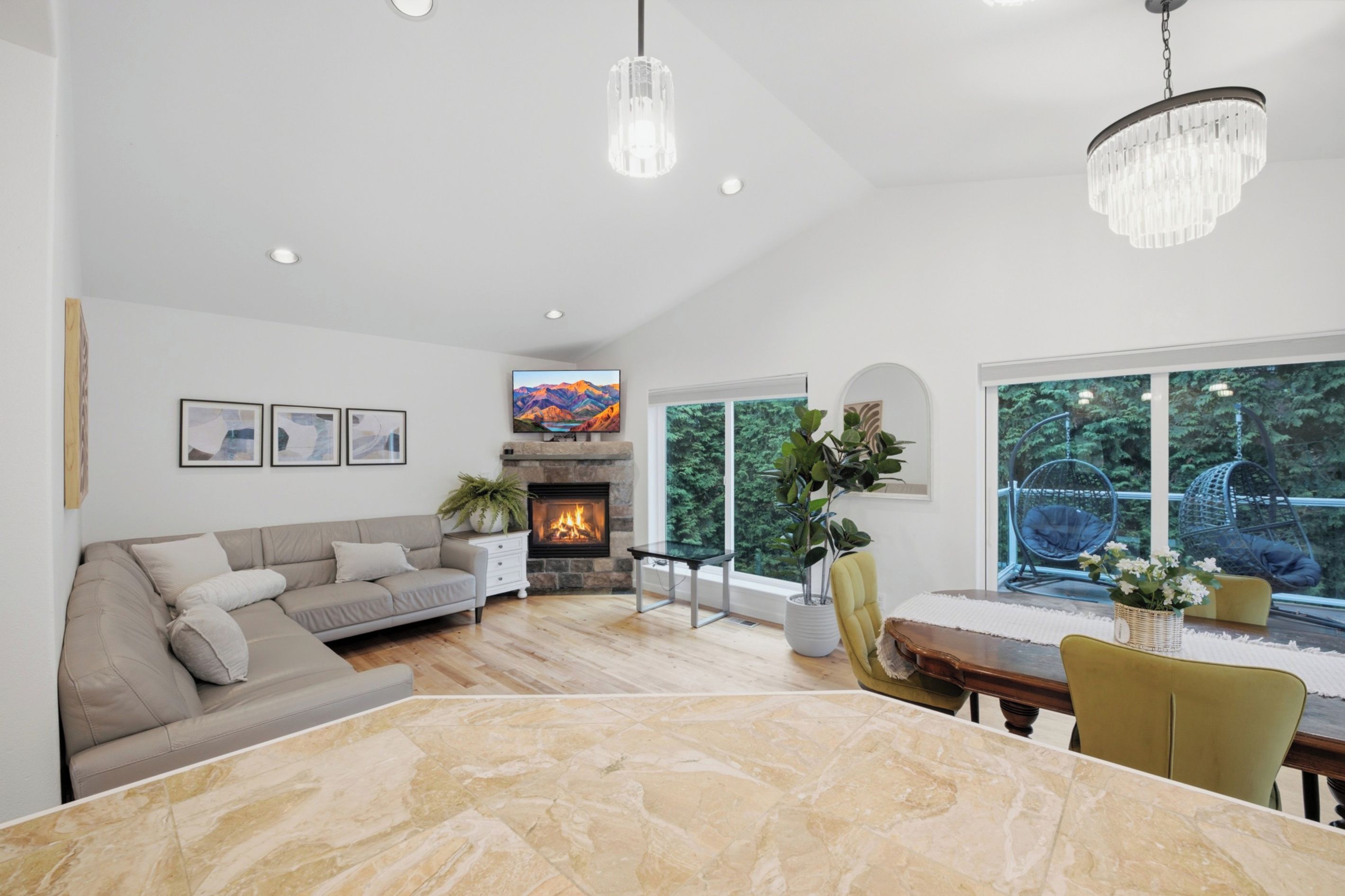 A spacious living room with a fireplace, a dining area with a marble table, and large windows offering a view of greenery.