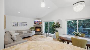 A spacious living room with a fireplace, a dining area with a marble table, and large windows offering a view of greenery.