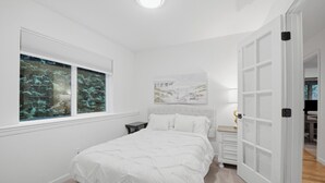 Een slaapkamer met een bed, een nachtkastje, een raam met uitzicht op bomen en een deur naar een andere kamer.