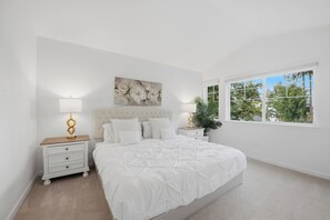 Ein Schlafzimmer mit einem großen Bett, weißen Nachttischen, einer Kopfteil, einem Gemälde, einem Fenster mit Blick auf Bäume und einer Topfpflanze.