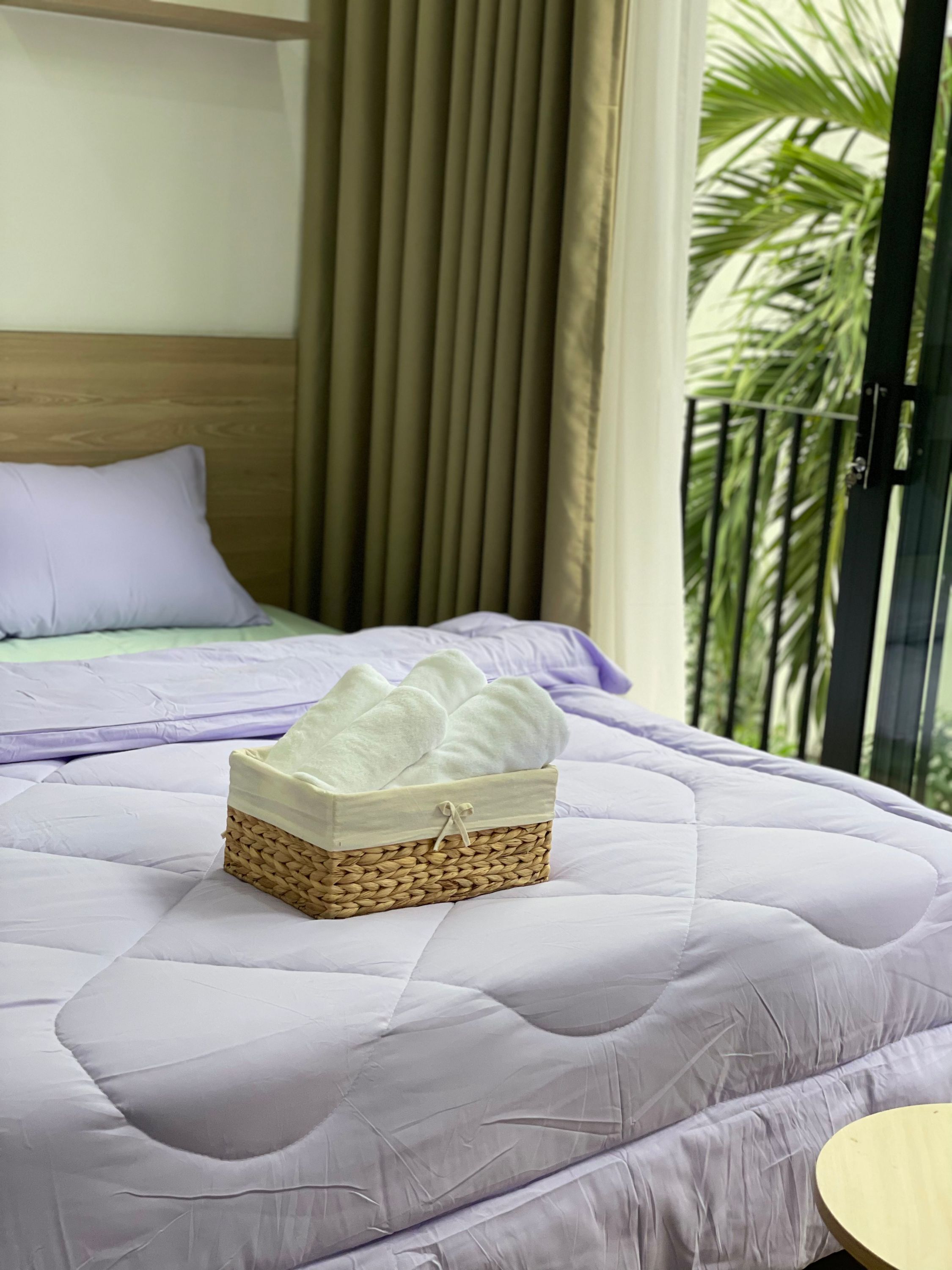 A bed with a purple quilt, a wicker basket with towels, and a view of palm trees through a window.