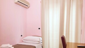 Uma cama de solteiro com colcha bege, uma escrivaninha de madeira, uma cadeira e uma janela com cortinas bege.