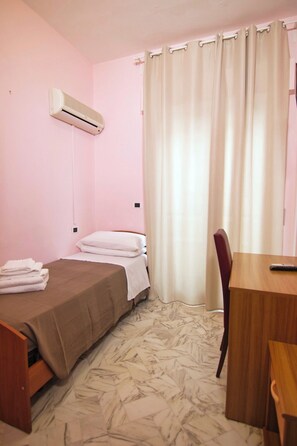 A single bed with a beige bedspread, a wooden desk, a chair, and a window with beige curtains. - HOTEL ROMA (Pineto)