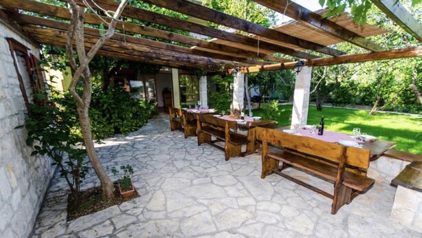 Un patio exterior con suelo de piedra, mesas y bancos de madera, y una estructura de pérgola.