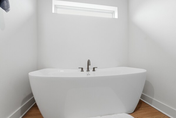 Een moderne badkamer met een vrijstaand bad, een raam boven de wastafel en een houten vloer.