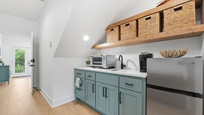 Eine moderne Küche mit türkisfarbenen Schränken, einem Edelstahlkühlschrank, einer Mikrowelle und einer Kaffeemaschine. Über der Arbeitsplatte befindet sich ein Holzregal mit gewebten Körben.