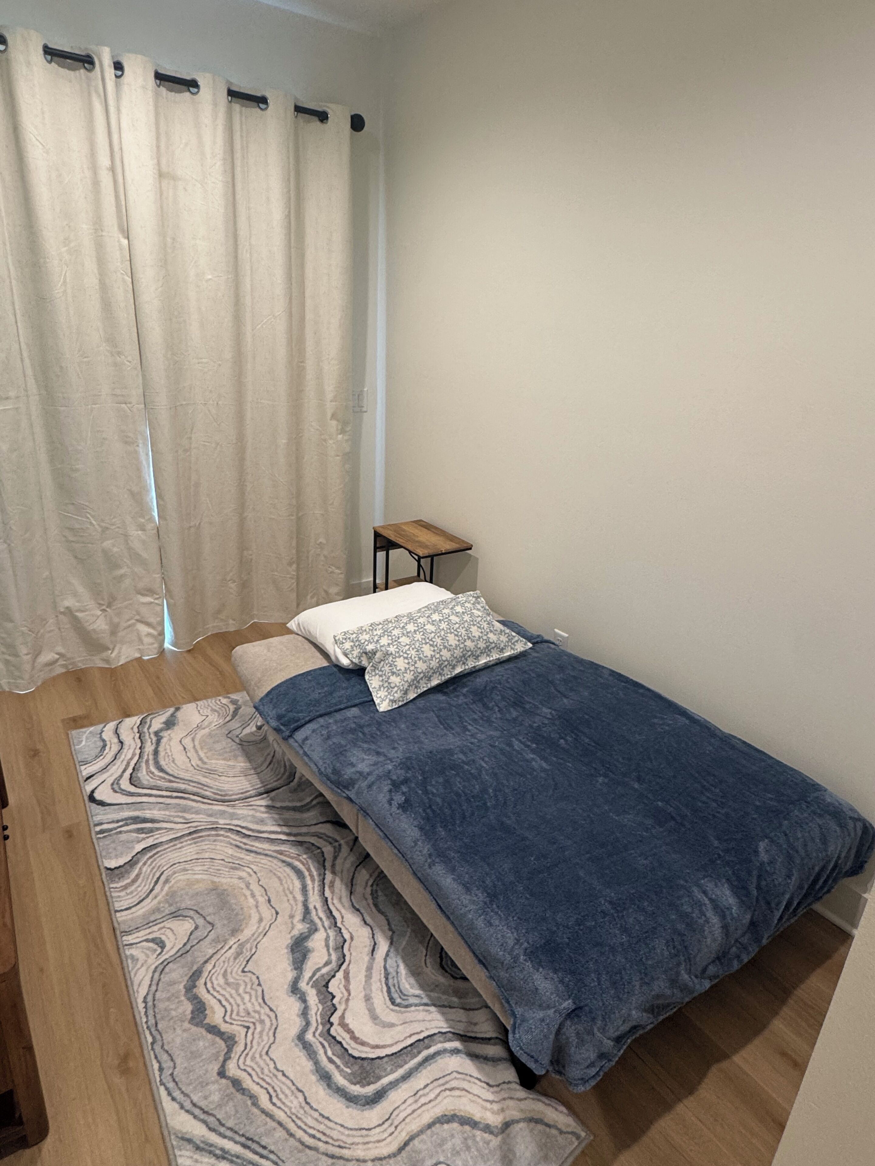 A small, single-bed room with a wooden floor, a blue bedspread, a patterned rug, a small bedside table, and a curtain.