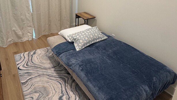 Ein kleines Zimmer mit einem Einzelbett, Holzboden, einer blauen Bettdecke, einem gemusterten Teppich, einem kleinen Nachttisch und einem Vorhang.