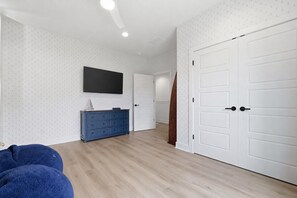 A room with a wooden floor, a blue dresser, a flat-screen TV mounted on the wall, and white doors. - Luxury Lakefront Escape on Beaver Lake - Sleeps the Whole Crew (Rogers)