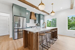 A modern kitchen with stainless steel appliances, a wooden island with bar stools, and a large window offering a view of greenery. - Luxury Lakefront Escape on Beaver Lake - Sleeps the Whole Crew (Rogers)