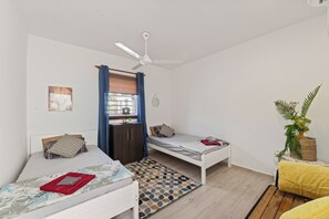 Een kamer met twee enkele bedden, een plafondventilator, een raam met blauwe gordijnen, een kleine plant en een spiegel aan de muur.