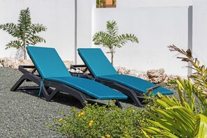 Twee lounge stoelen met blauwe kussens, geplaatst op een grindpad, met een witte muur en kleine bomen op de achtergrond.