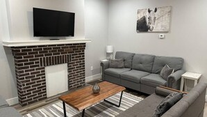 Una sala de estar con chimenea, un televisor de pantalla plana montado encima, un sofá gris, una mesa de centro de madera y una alfombra a rayas.