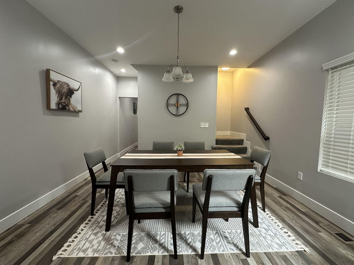 A dining area with a rectangular table, six chairs, a clock on the wall, and a staircase leading to an upper level.