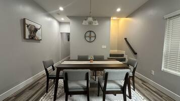 A dining area with a rectangular table, six chairs, a clock on the wall, and a staircase leading to an upper level.