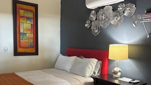 Un dormitorio con cama, mesita de noche, lámpara, aire acondicionado instalado en la pared y un adorno en la pared.
